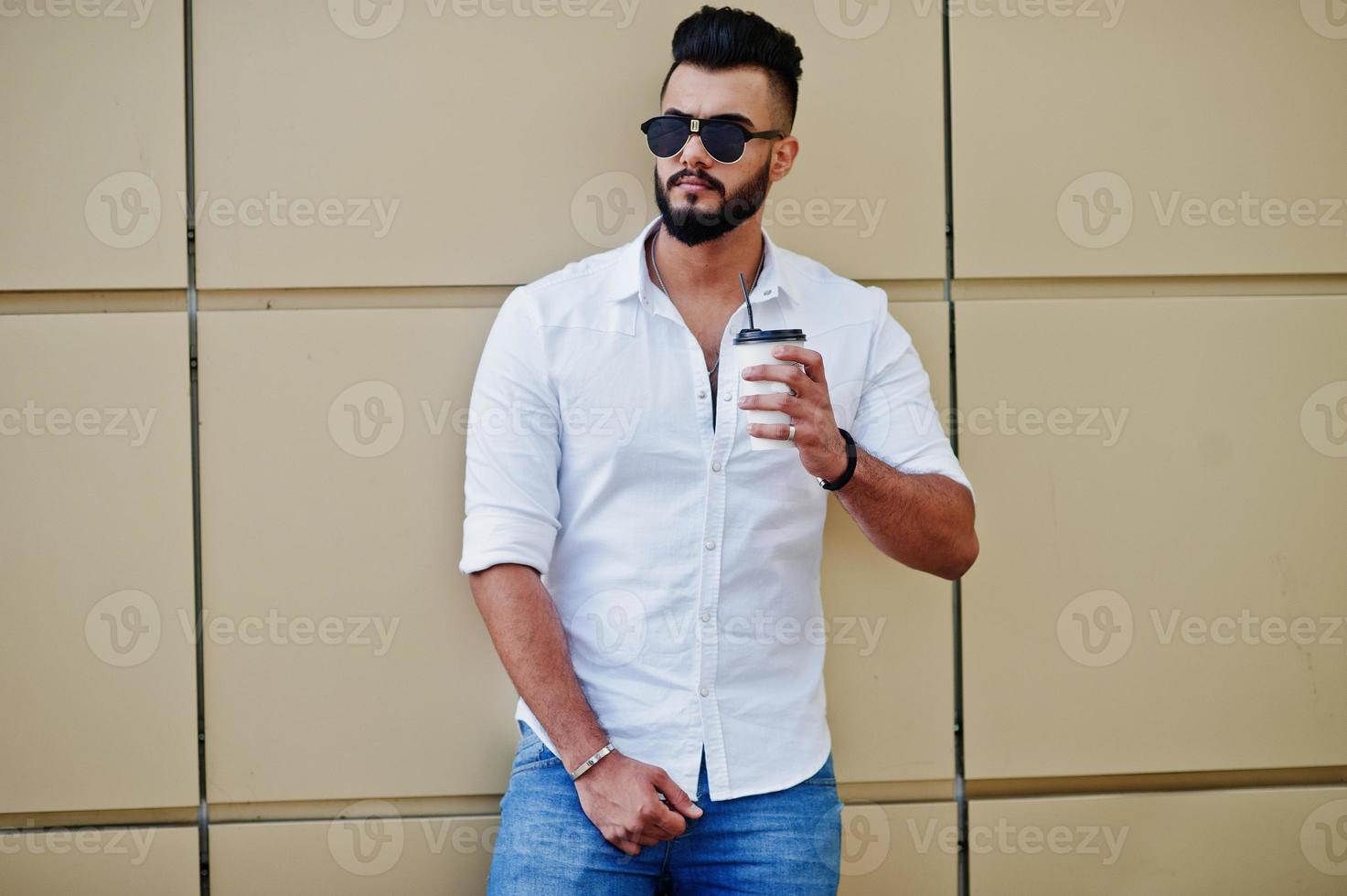 Stylish tall arabian man model in white shirt, jeans and sunglasses posed at street of city. Beard attractive arab guy with cup of coffee against golden wall. photo