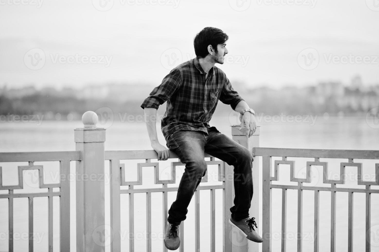 Young indian student man at checkered shirt and jeans sitting on handrails against lake. photo