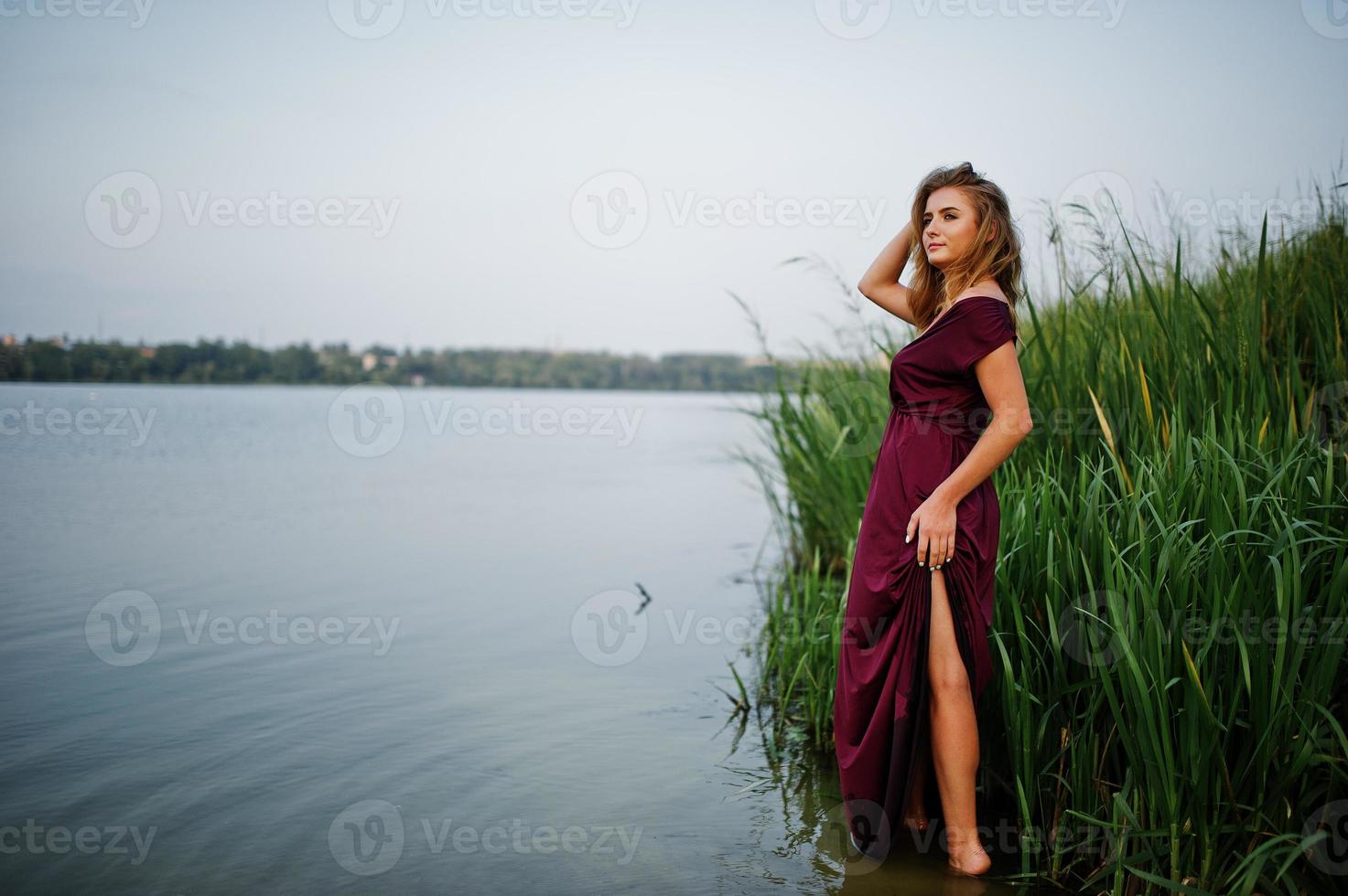 mujer sensual rubia en vestido rojo marsala de pie en el agua del lago con juncos. foto