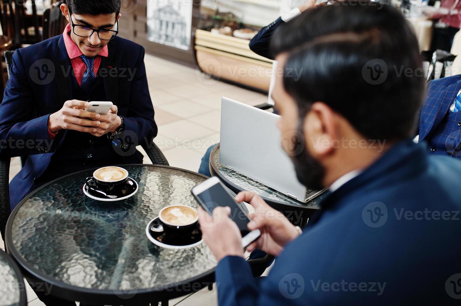 grupo de hombres de negocios indios en trajes sentados en la oficina en un café con una computadora portátil, enviando mensajes de texto por teléfono y haciendo fotos de café.