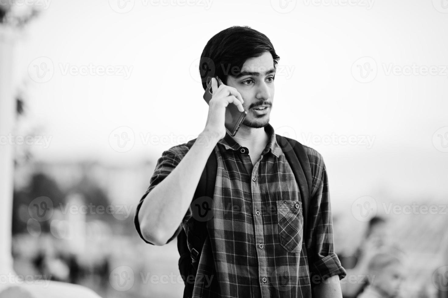 Young indian student man at checkered shirt and jeans with backpack speaking on mobile phone. photo