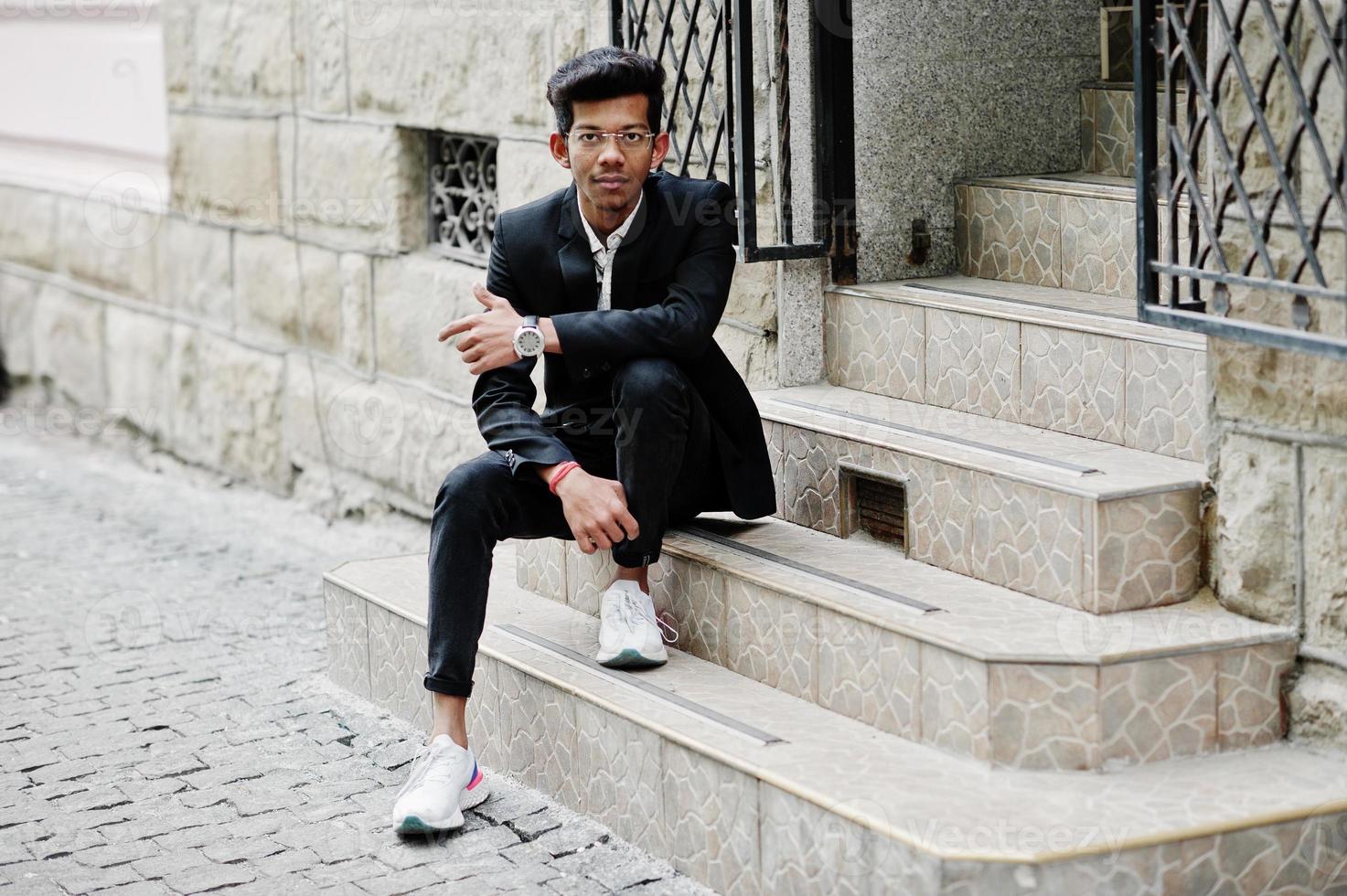 Young indian man on suit and glasses posed outdoor and sitting on stairs. photo