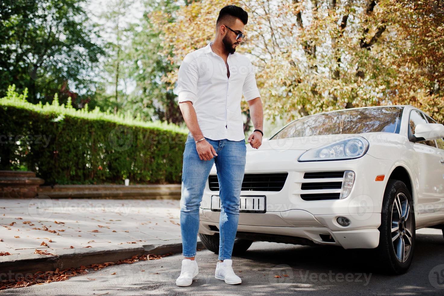 Stylish tall arabian man model in white shirt, jeans and sunglasses posed at street of city. Beard rich attractive arab guy against white suv car. photo
