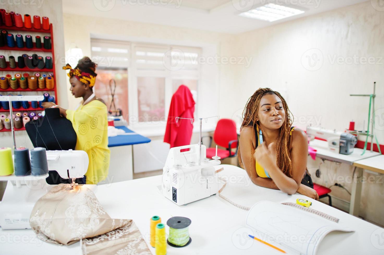 Two african dressmaker woman sews clothes on sewing machine at tailor office. Black seamstress girls. photo