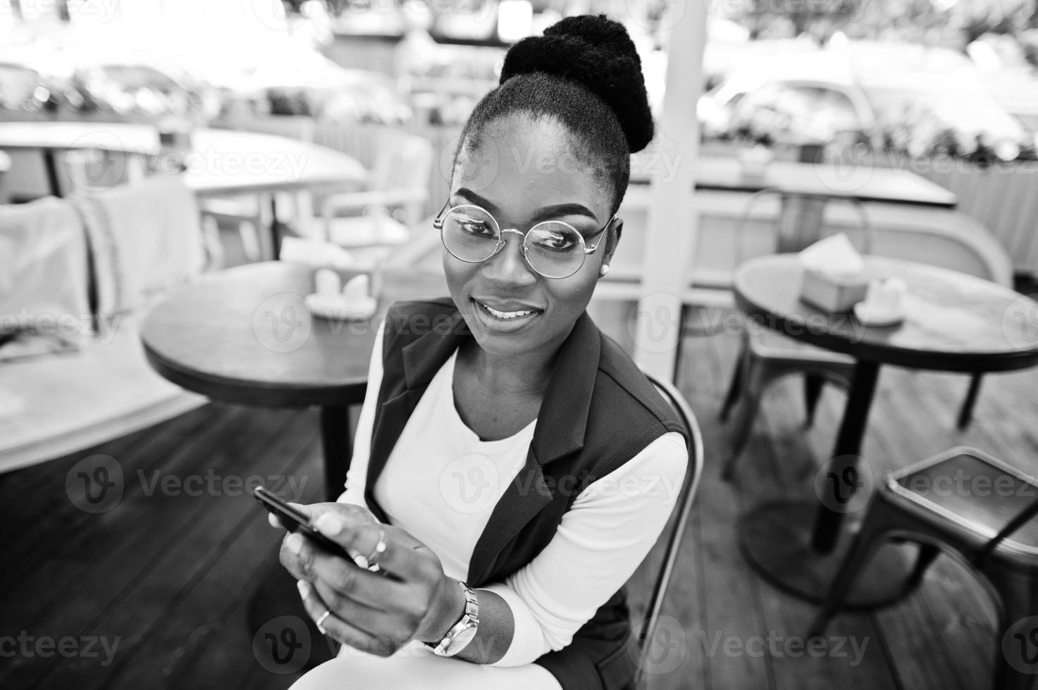 African american girl wear in glasses with mobile phone sitting at outdoor caffe. photo