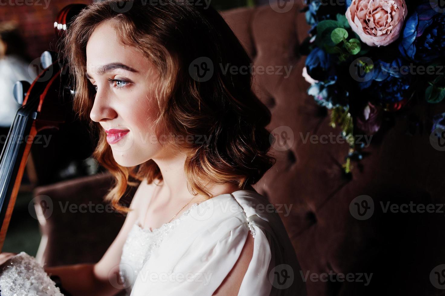 Pretty young gilrl musician in white dress with double bass sitting on brown vintage sofa. photo