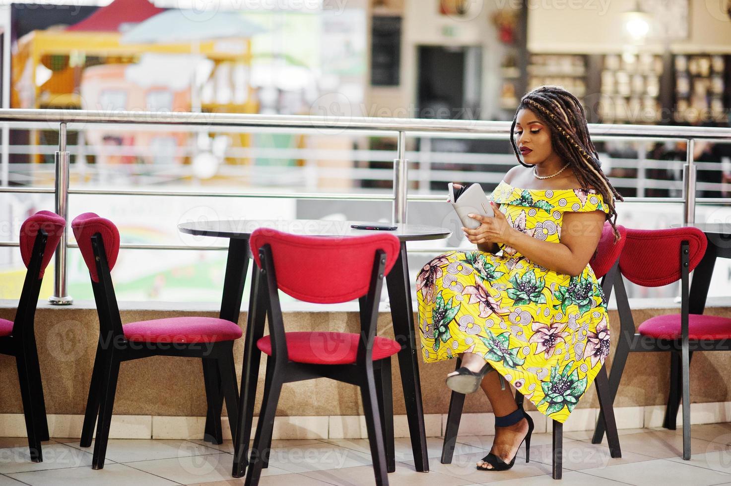 Cute small height african american girl with dreadlocks, wear at coloured yellow dress, sitting on cafe at shopping center and looking at her wallet. photo