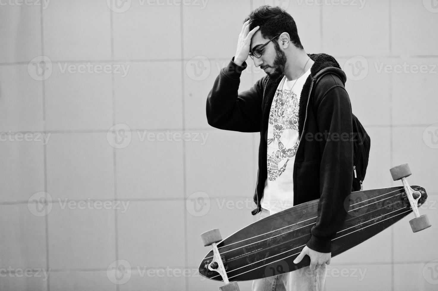 hombre árabe de estilo callejero en anteojos con longboard posado contra la pared gris. foto