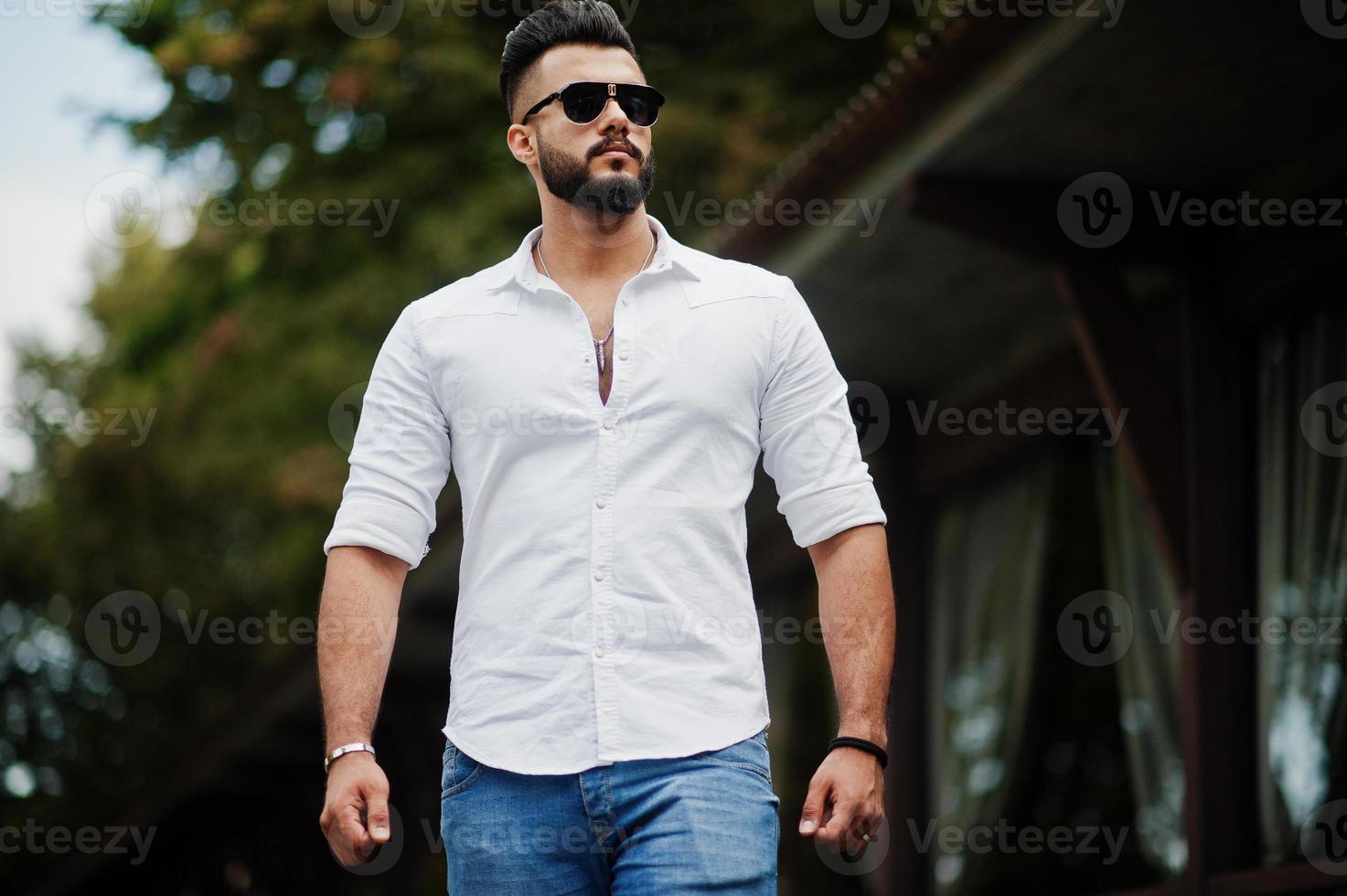 Stylish tall arabian man model in white shirt, jeans and sunglasses walking at street of city. Beard attractive arab guy. photo