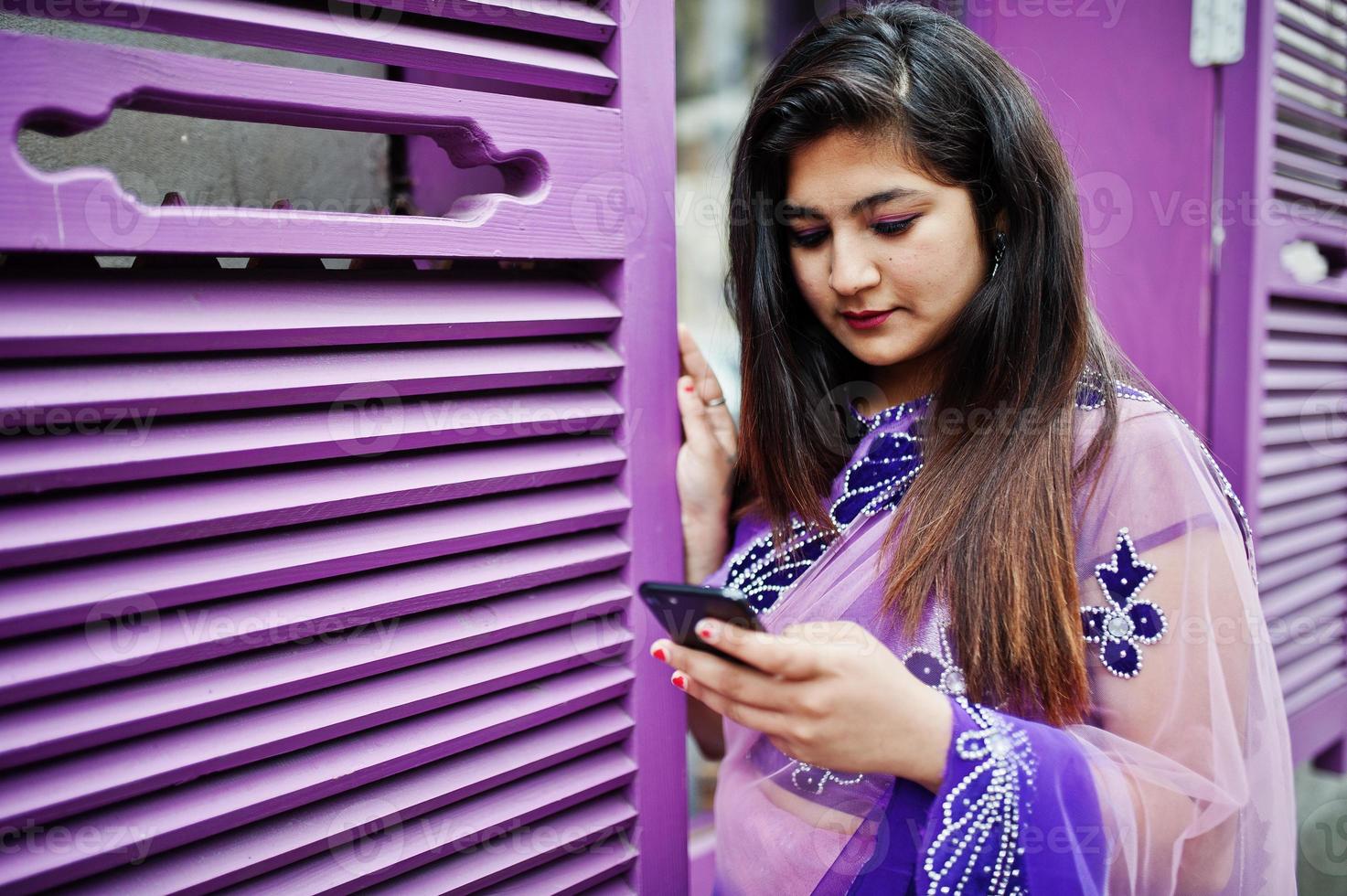 Indian hindu girl at traditional violet saree posed at street against purple windows with mobile phone at hands. photo