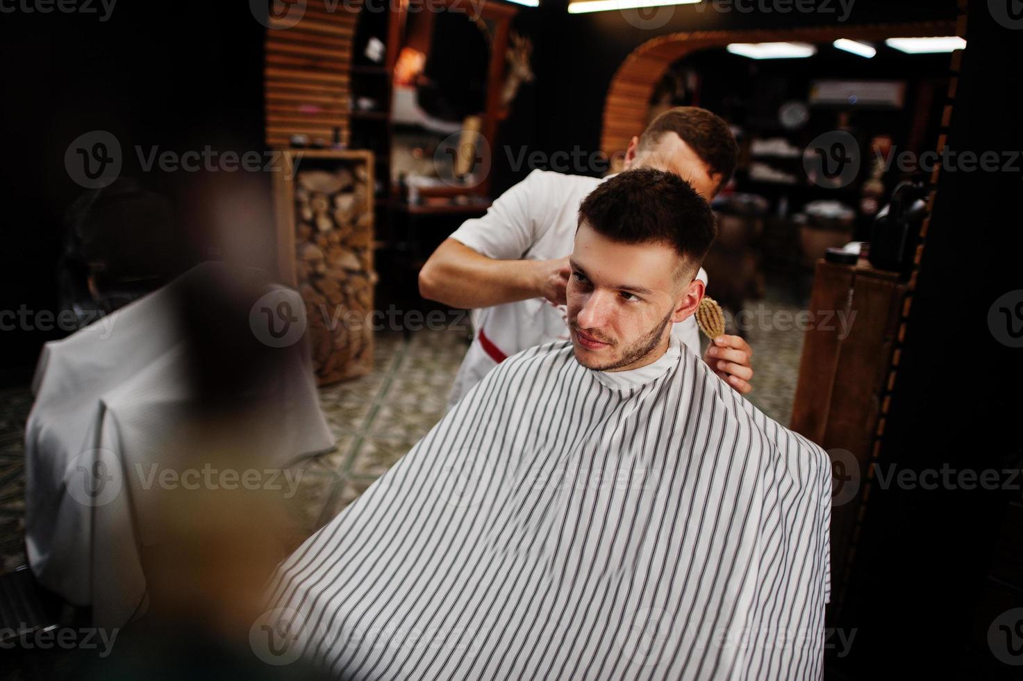 Young bearded man getting haircut by hairdresser while sitting in chair at barbershop. Barber soul. photo