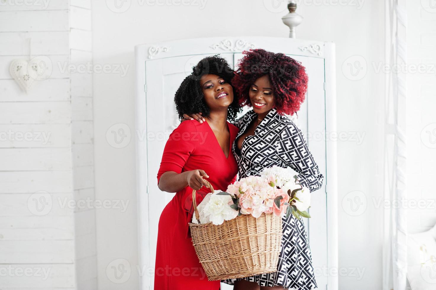 dos mujeres afroamericanas de moda vestidas de noche de pie con una cesta de flores en las manos contra el viejo armario vintage en la habitación blanca. foto