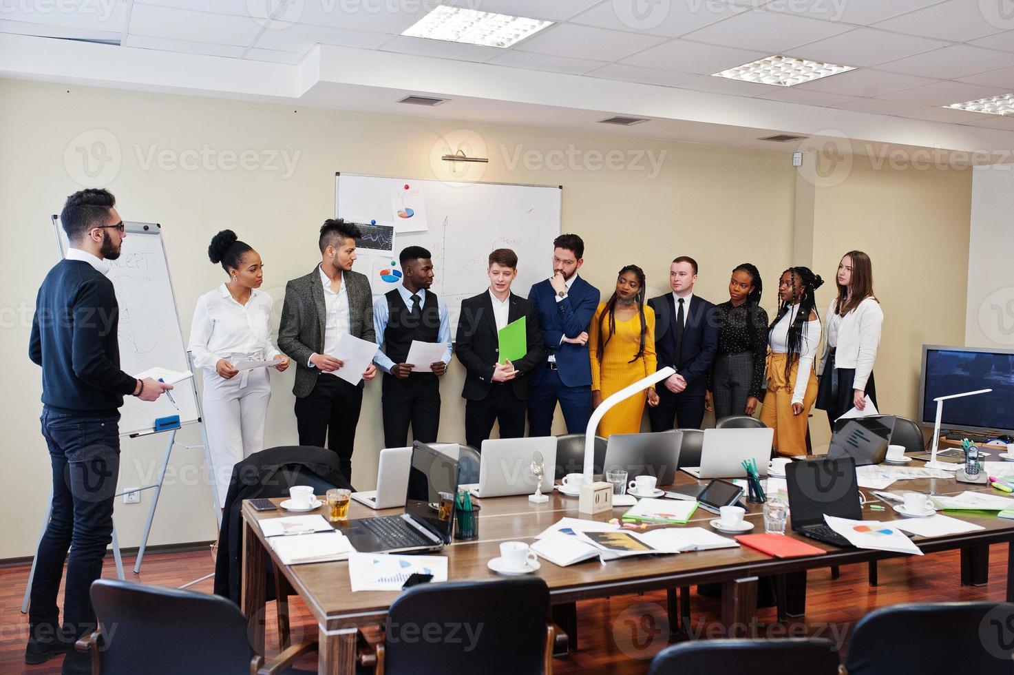 Mixed race business coach presenting report standing near whiteboard pointing on sales statistic shown on diagram and chart teach diverse company members gathered together in conference room. photo