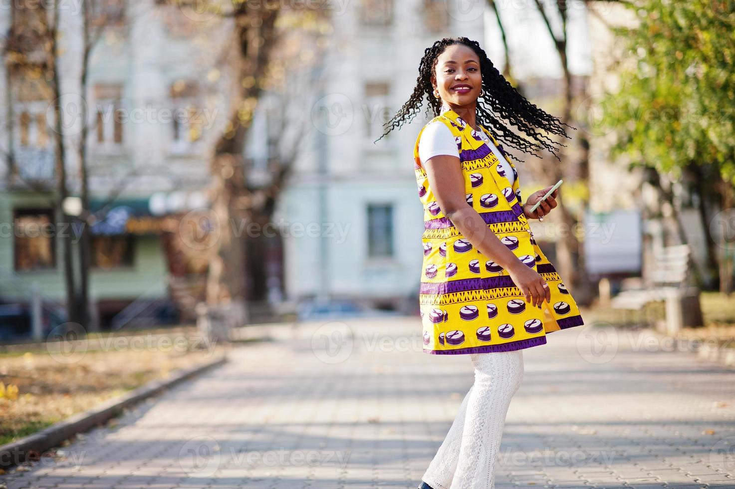Stylish african american women in yellow jacket posed on street at sunny day with mobile phone at hand. photo