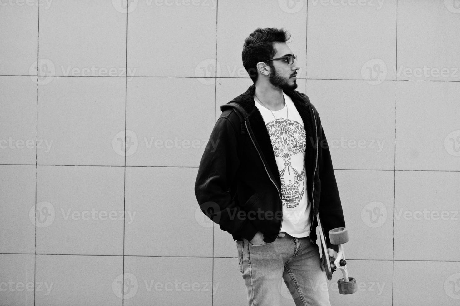 Street style arab man in eyeglasses with longboard posed against gray wall. photo