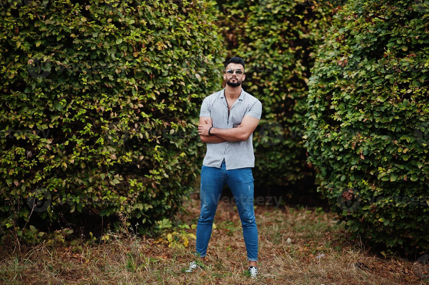 el hombre de barba árabe alto de moda usa camisa, jeans y gafas de sol posados en el parque contra la vegetación. foto