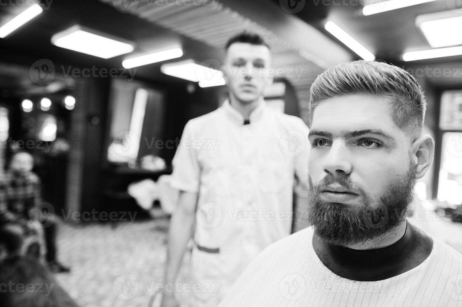 Young bearded man getting haircut by hairdresser while sitting in chair at barbershop. Barber soul. photo