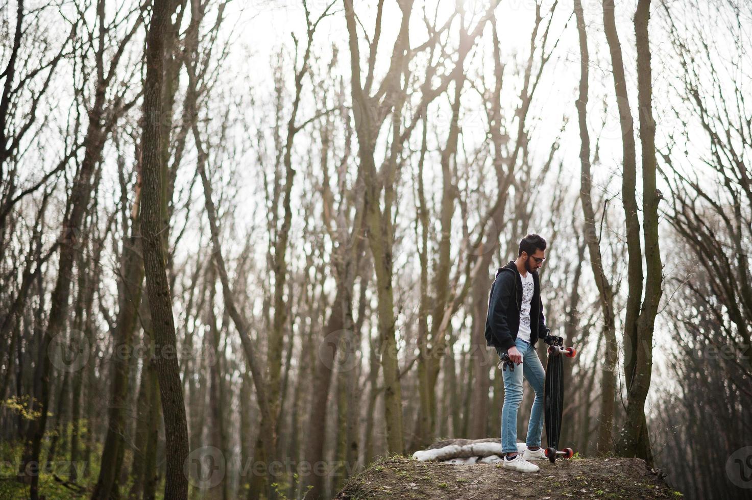 Street style arab man in eyeglasses with longboard on wood. photo