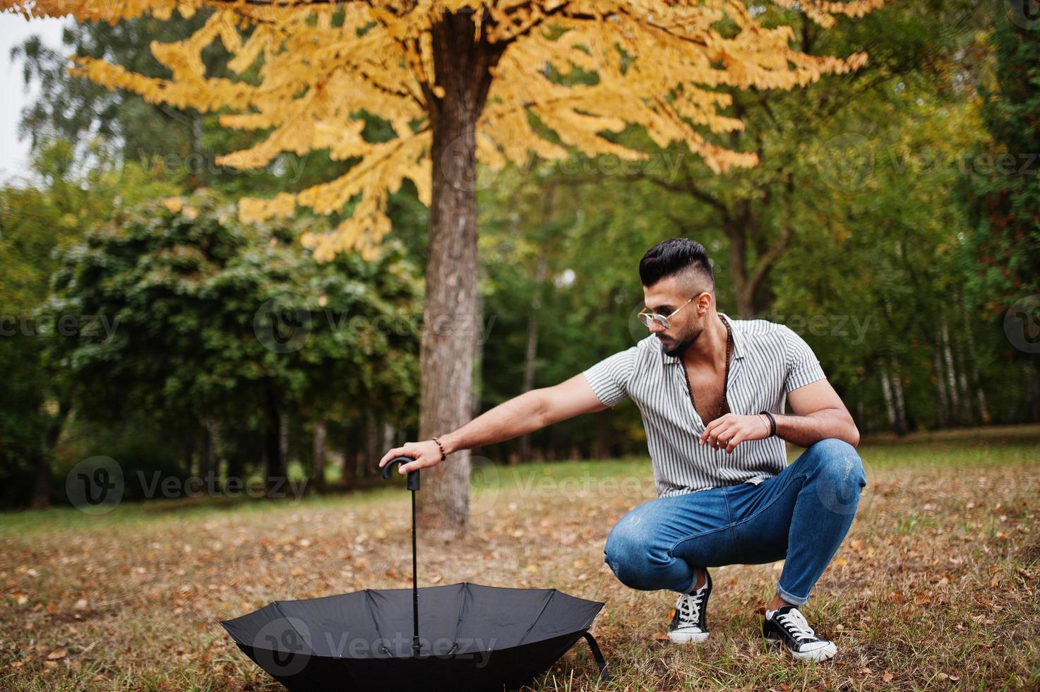 Fashionable tall arab beard man wear on shirt, jeans and sunglasses posed on autumn park with umbrella against yellow leaves tree. photo