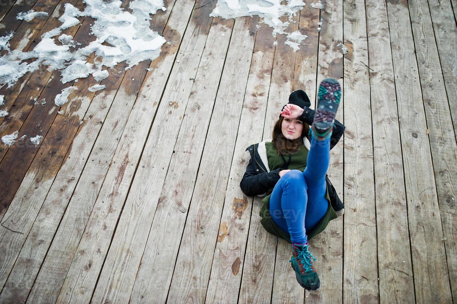 Young girl wear on long green sweatshirt, jeans and black headwear sitting on wooden floor with snow. photo