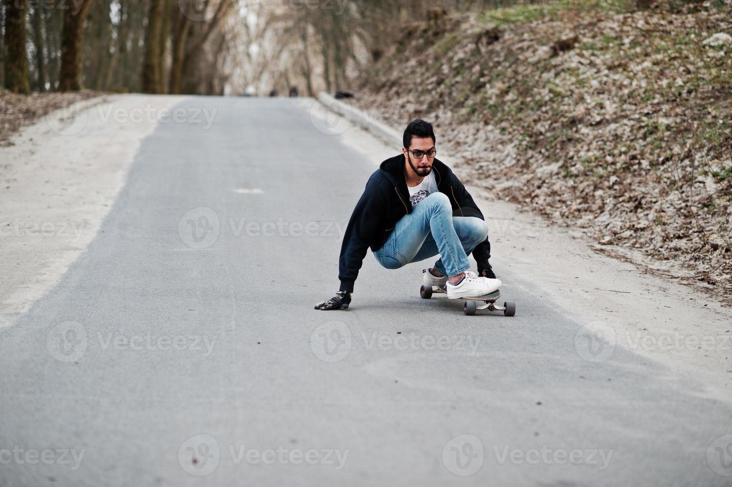 Street style arab man in eyeglasses with longboard longboarding down the road. photo