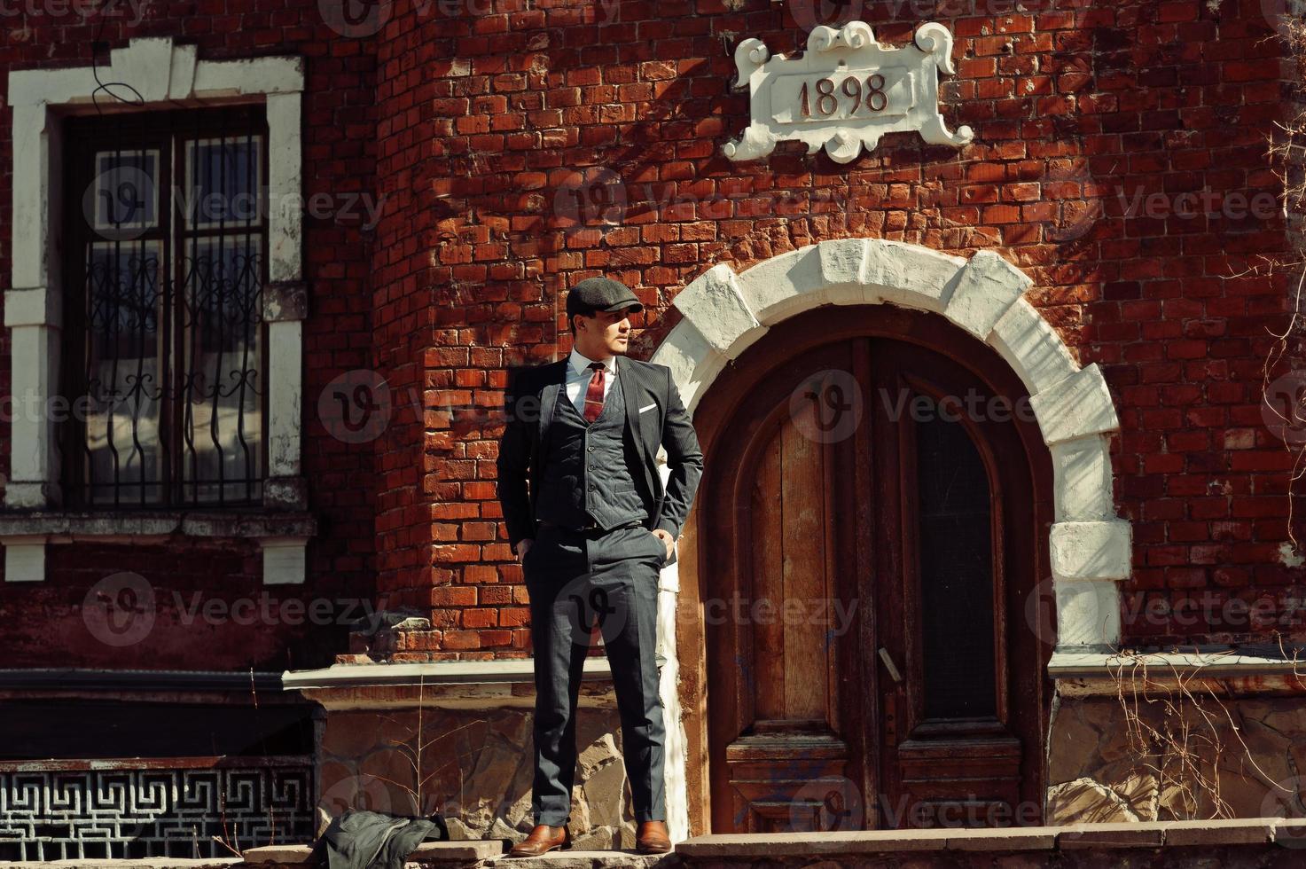 Portrait of retro 1920s english arabian business man wearing dark suit, tie and flat cap standing against old brick house 1898 year. photo