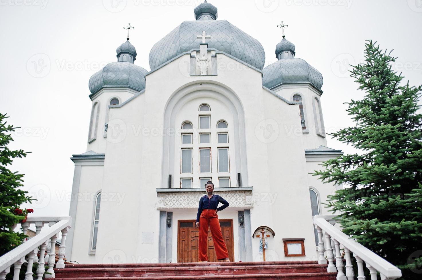 mujer africana con pantalones naranjas y camisa azul posada contra una gran iglesia. fe y creer en dios. foto