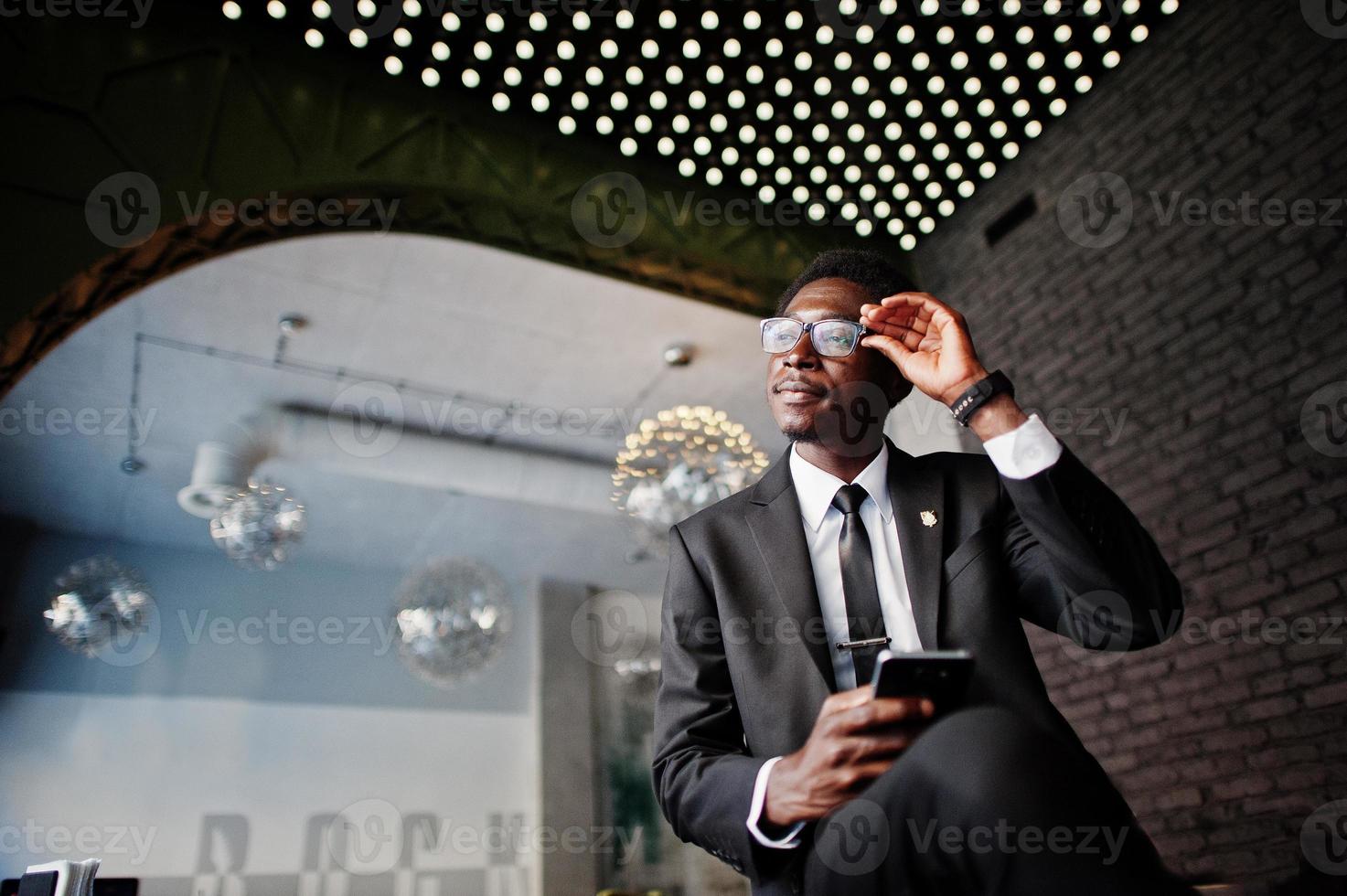 el hombre afroamericano de negocios usa traje negro y anteojos en la oficina mirando el teléfono móvil contra luces futuristas. foto