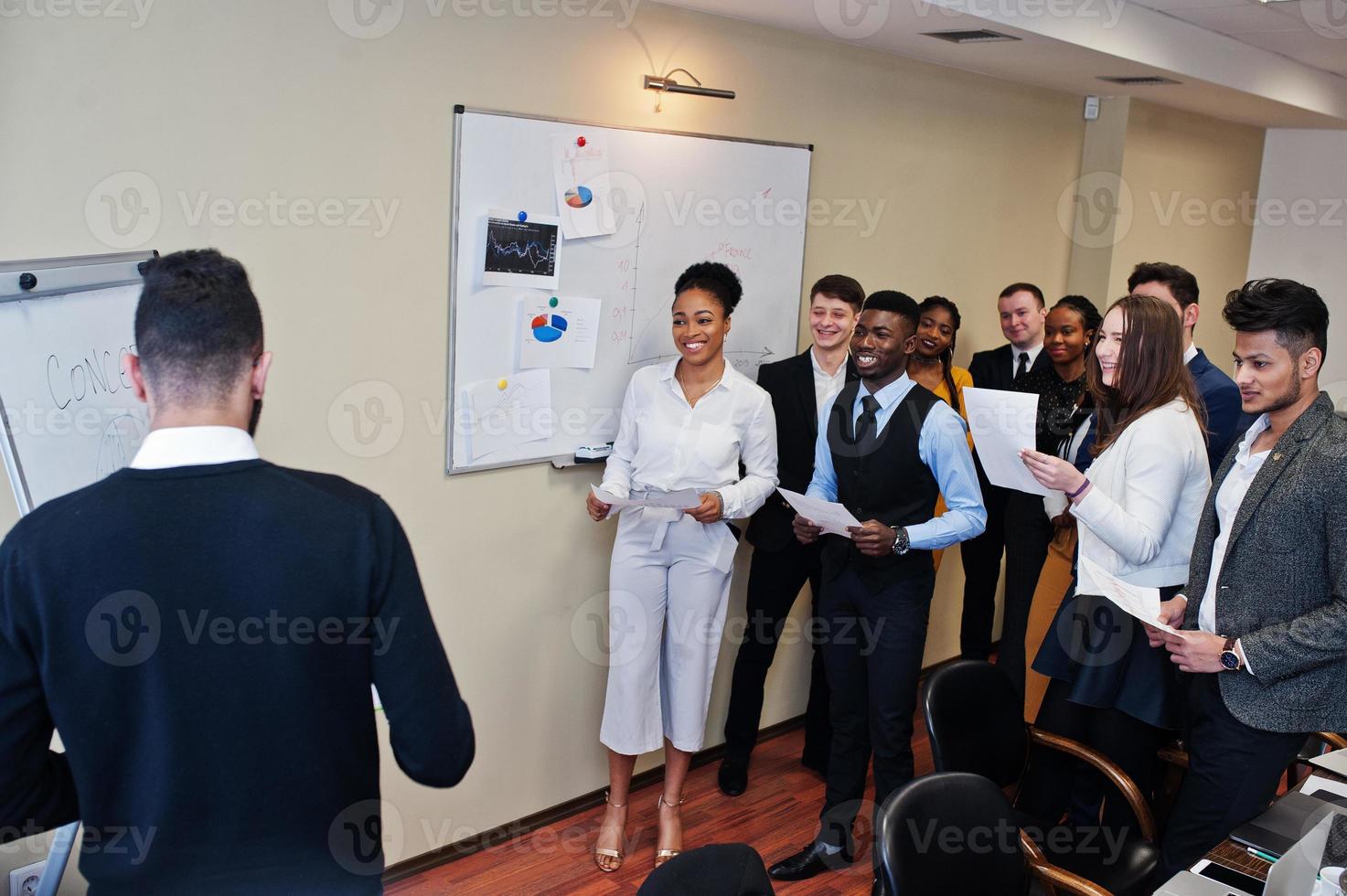 Mixed race business coach presenting report standing near whiteboard pointing on sales statistic shown on diagram and chart teach diverse company members gathered together in conference room. photo