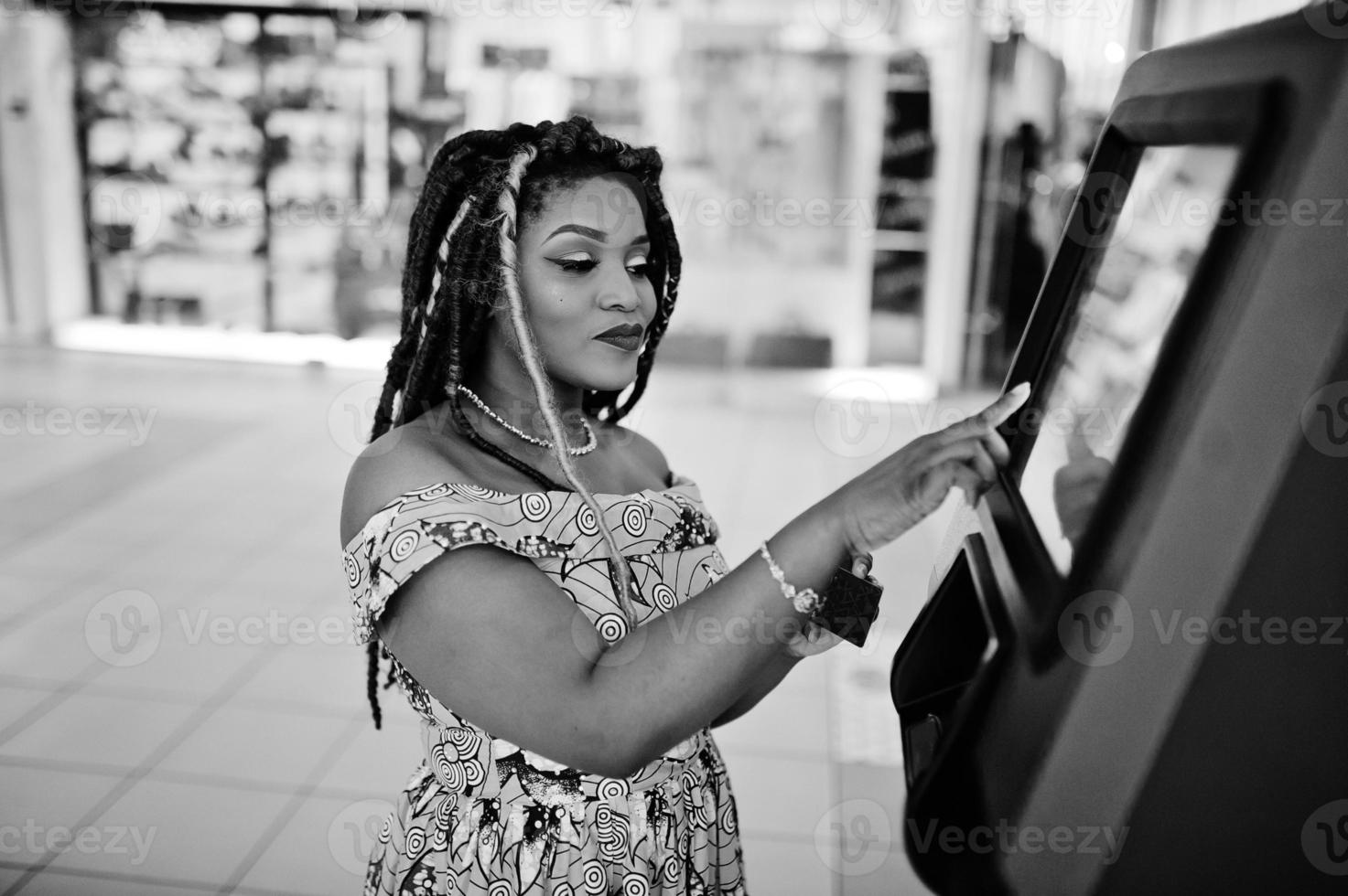 linda chica afroamericana de pequeña altura con rastas, vestida con un vestido amarillo de color, contra un cajero automático con tarjeta de crédito a mano. en blanco y negro. foto