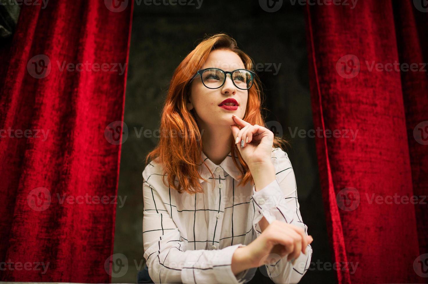 Attractive redhaired woman in eyeglasses, wear on white blouse posing at arch of open red curtains. photo