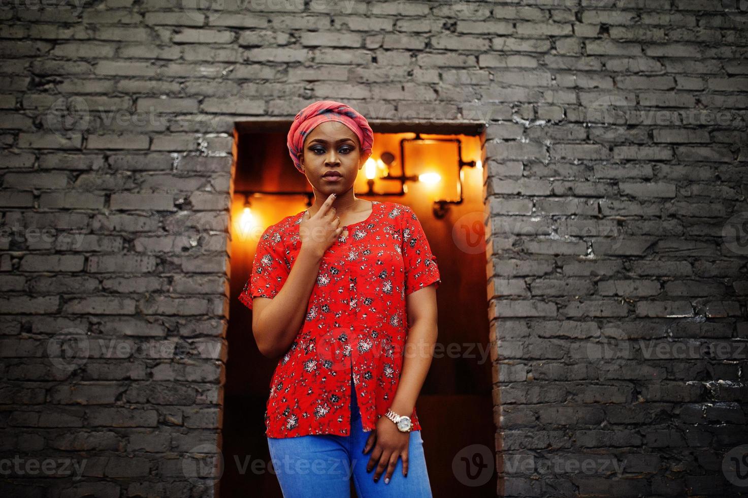 Stylish african woman in red shirt and hat posed against brick wall. photo