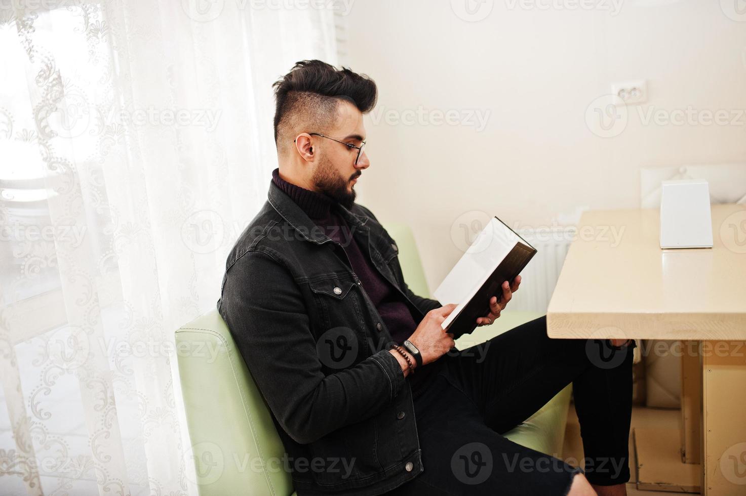 Arab man wear on black jeans jacket and eyeglasses sitting in cafe, read book. Stylish and fashionable arabian model guy. photo