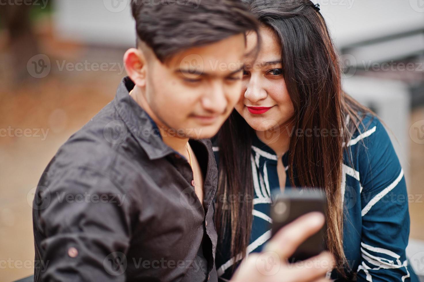 pareja india posó al aire libre, sentada en un banco juntos y mirando el teléfono móvil, haciendo selfie. foto