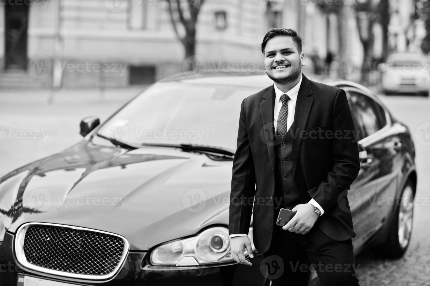 Stylish indian businessman in formal wear with mobile phone standing against black business car on street of city. photo