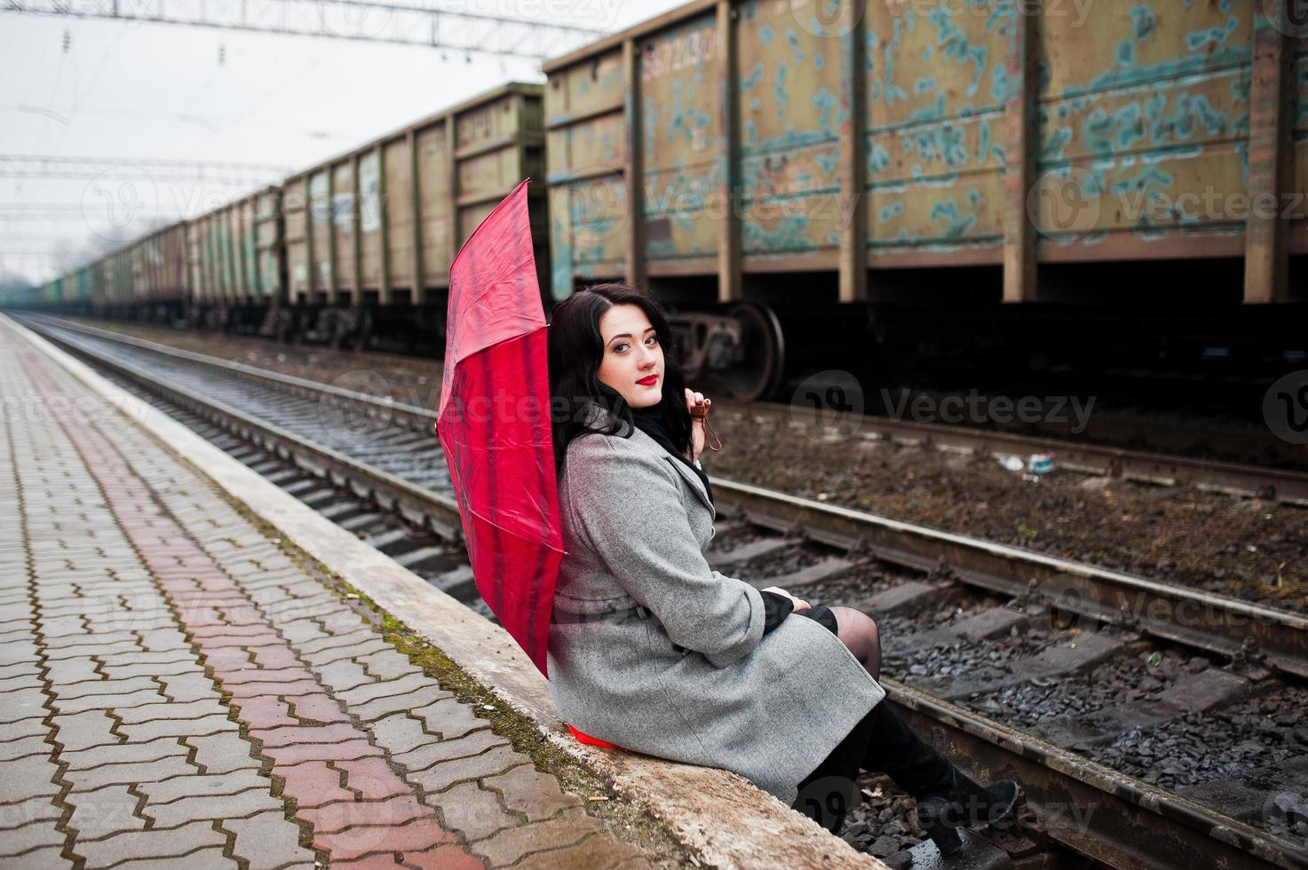 chica morena con abrigo gris con paraguas rojo en la estación de tren. foto