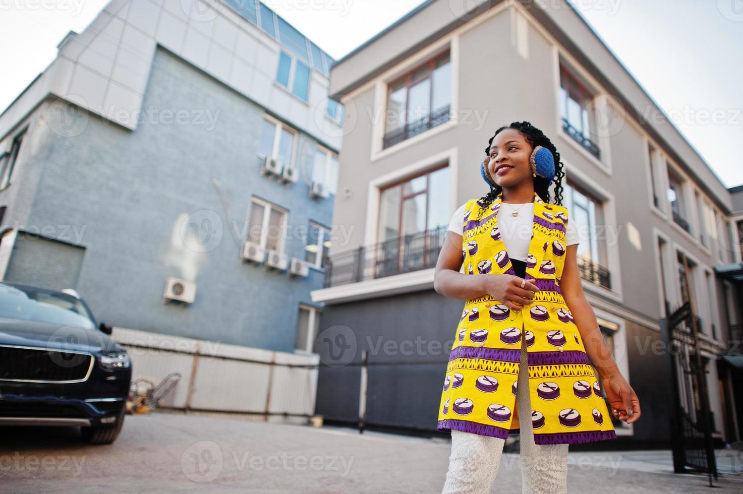 Stylish african american women in yellow jacket posed and earphones on street against modern building. photo