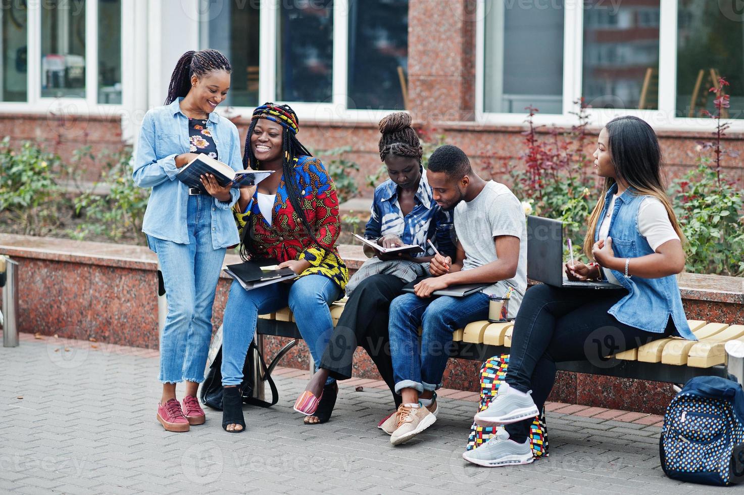 Group of five african college students spending time together on campus at university yard. Black afro friends studying at bench with school items, laptops notebooks. photo