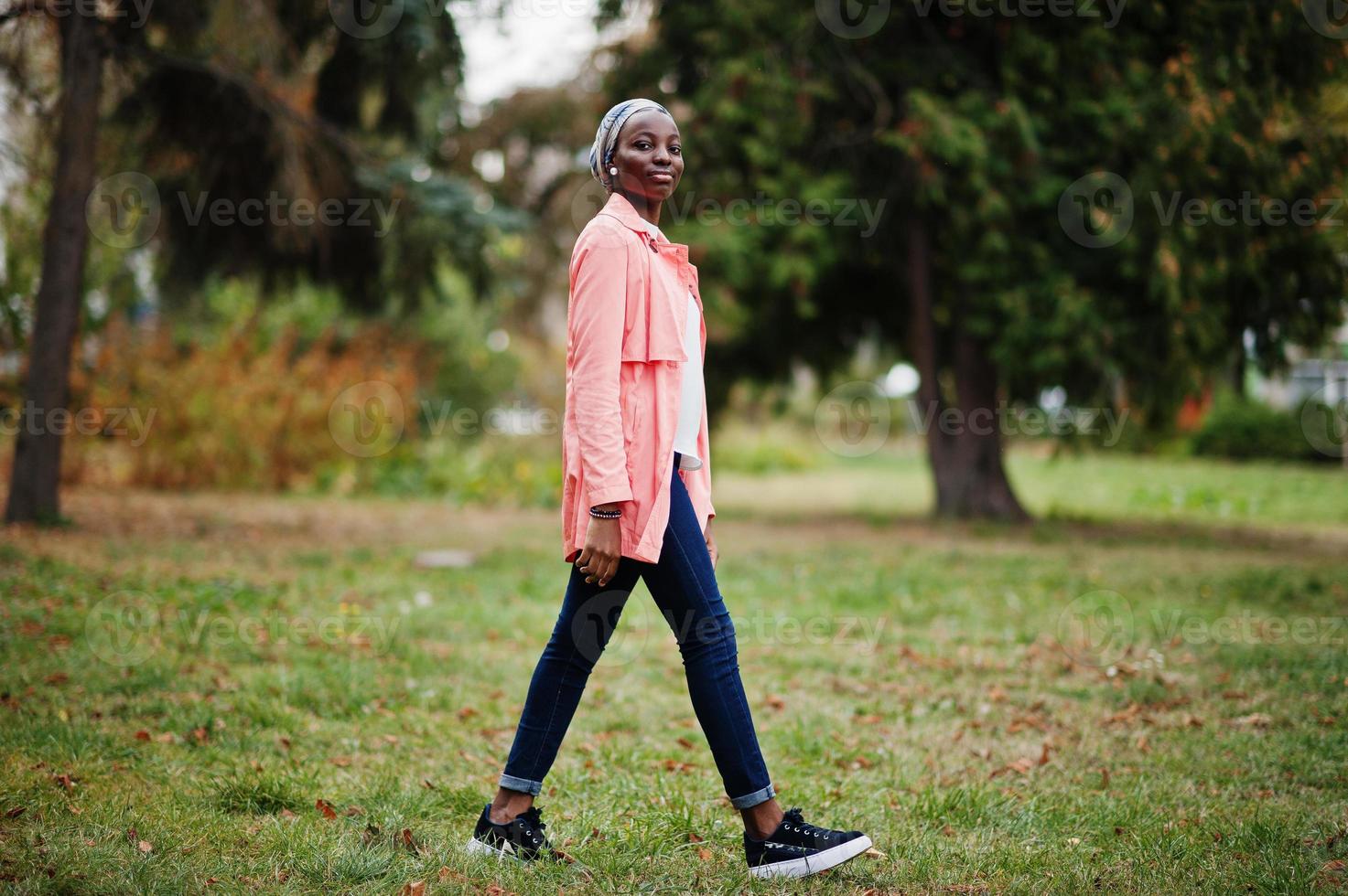 Young modern fashionable, attractive, tall and slim african muslim woman in hijab or turban head scarf and pink coat posed at park. photo