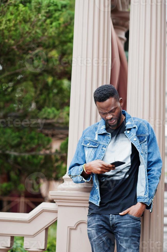 Young attractive serious african american man model. Modern handsome guy in jeans jacket standing on urban city street and looking at his mobile phone. photo