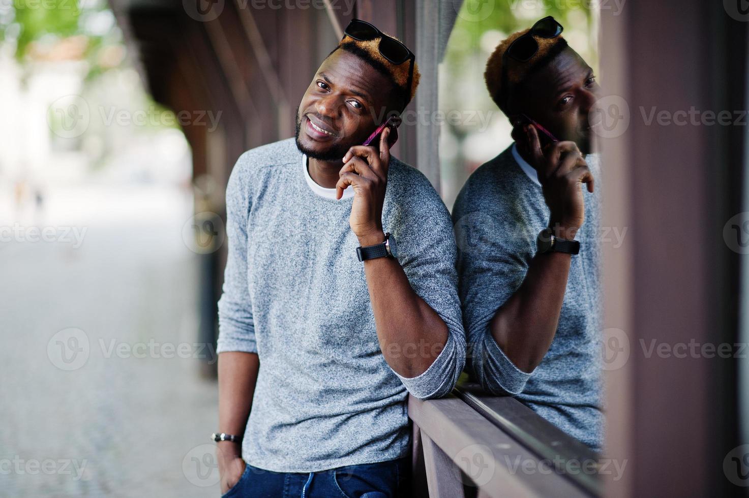 elegante chico afroamericano con suéter gris y gafas de sol negras posado en la calle y hablando por teléfono. chico negro de moda. foto