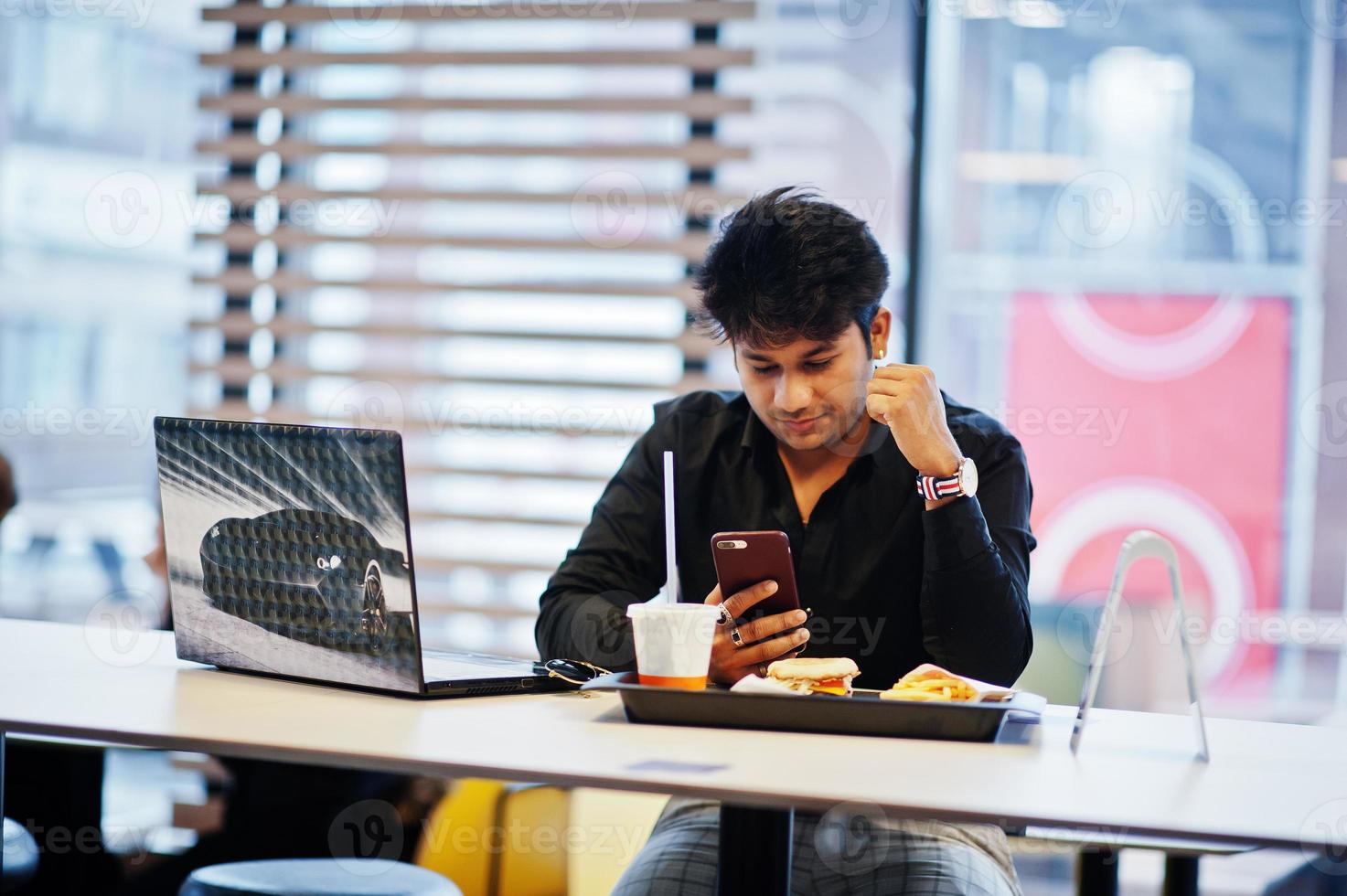 Stylish indian man sitting at fast food cafe against his laptop with mobile phone at hand. photo