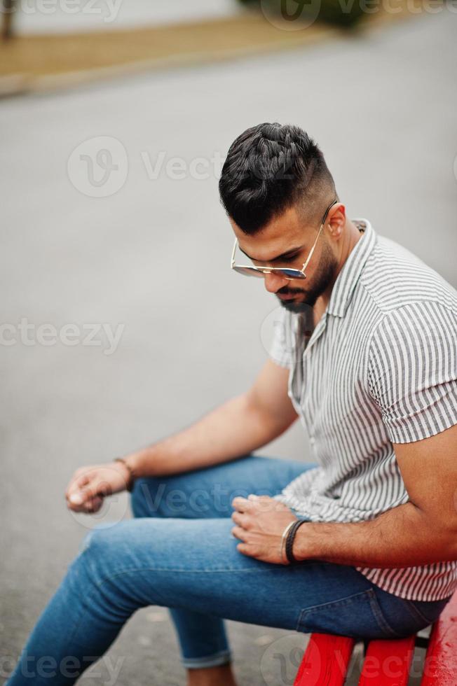 el hombre de barba árabe alto de moda usa camisa, jeans y gafas de sol sentado en un banco rojo en el parque. foto