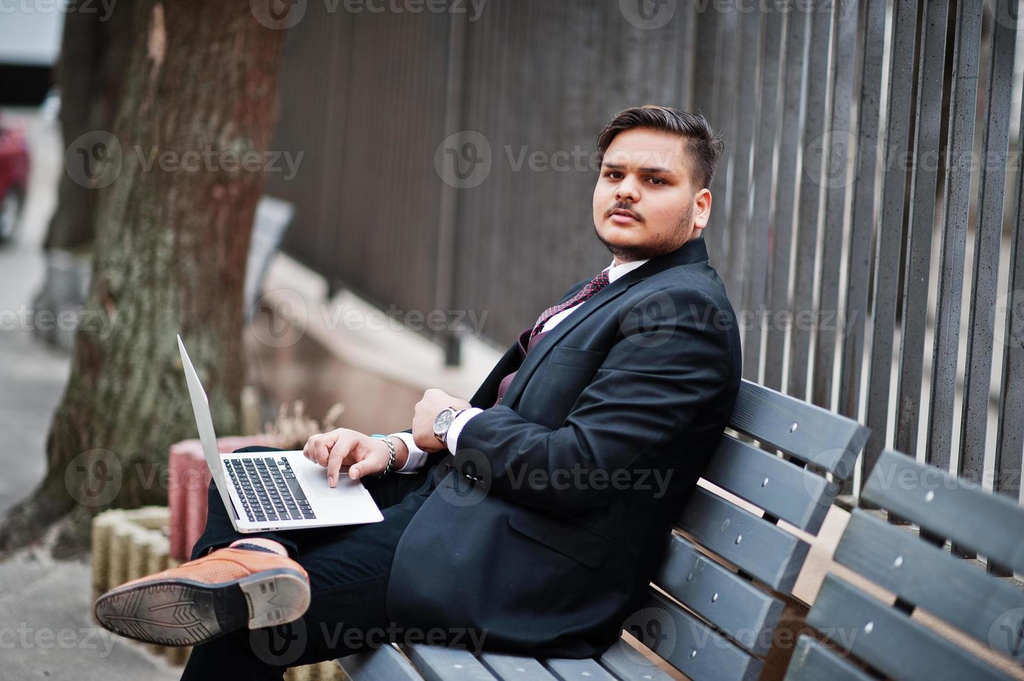 Stylish indian businessman in formal wear sitting on bench with laptop. photo