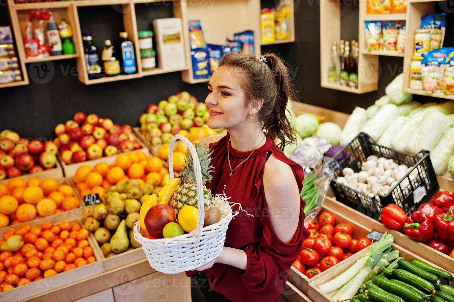 Girl in red holding different fruit and vegetables at basket on fruits store. photo