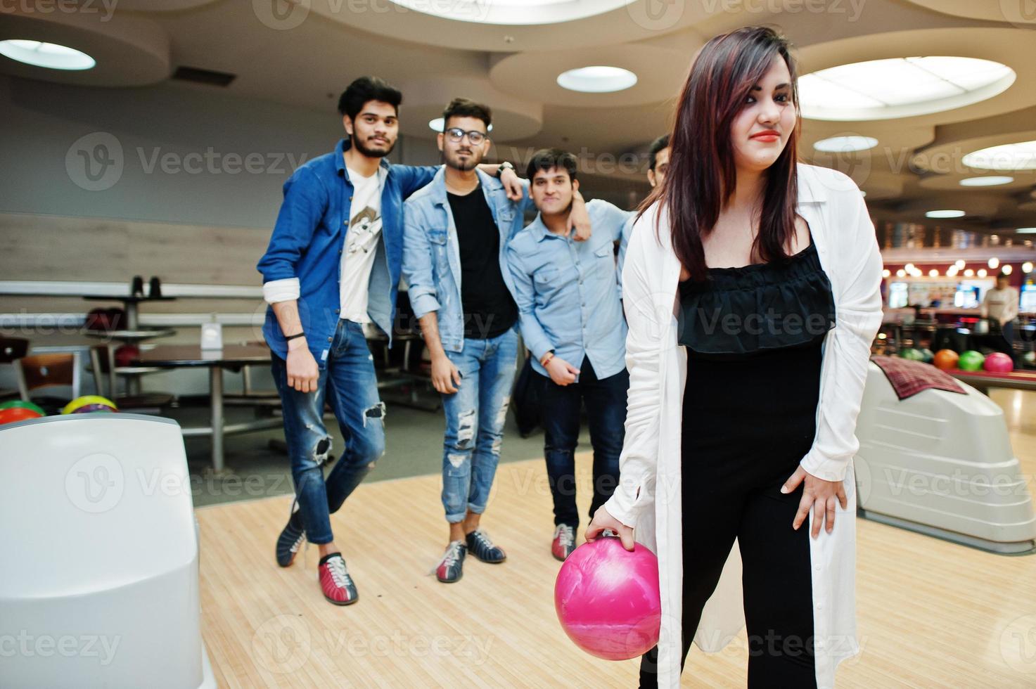 mujer del sur de asia parada en una bolera con pelota en las manos. la chica se está preparando para un lanzamiento. los amigos la apoyan en voz alta. foto