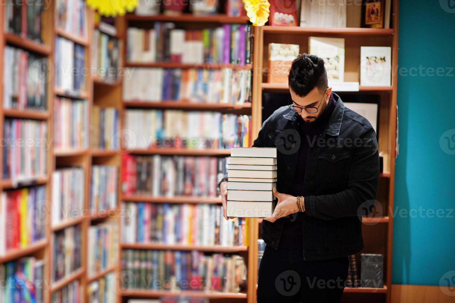 Tall smart arab student man, wear on black jeans jacket and eyeglasses, at library with stack of books. photo