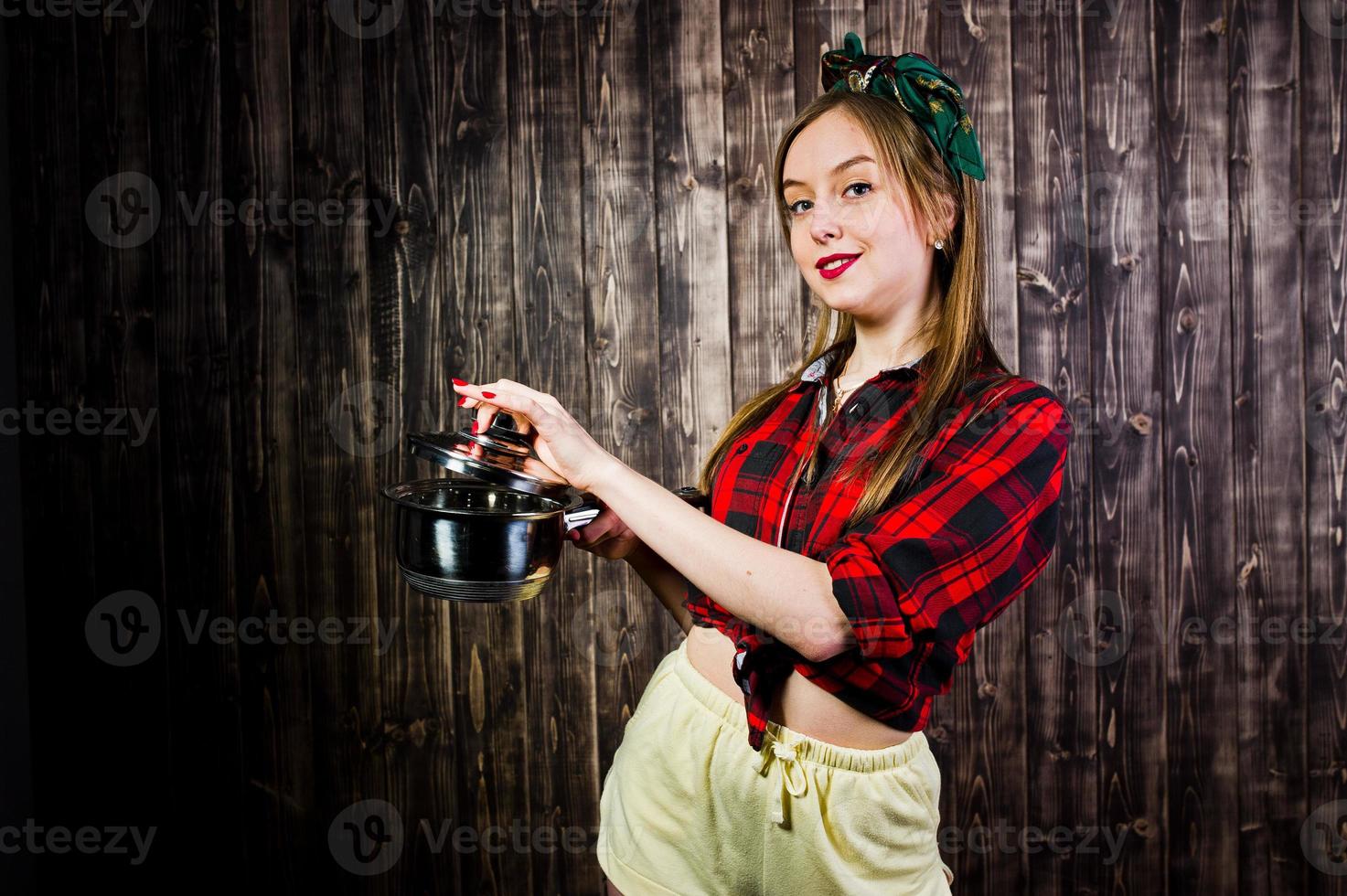 Young funny housewife in checkered shirt and yellow shorts pin up style with saucepan on wooden background. photo