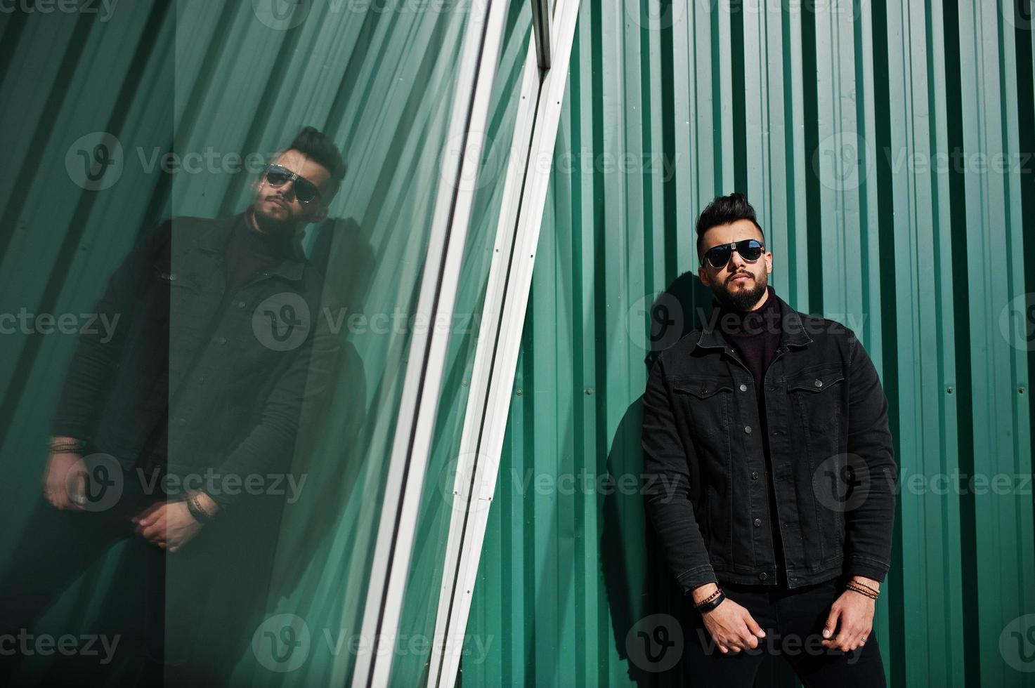 Fashion rich beard Arab man wear on black jeans jacket and sunglasses posed against green wall with windows. Stylish, succesful and fashionable arabian model guy. photo