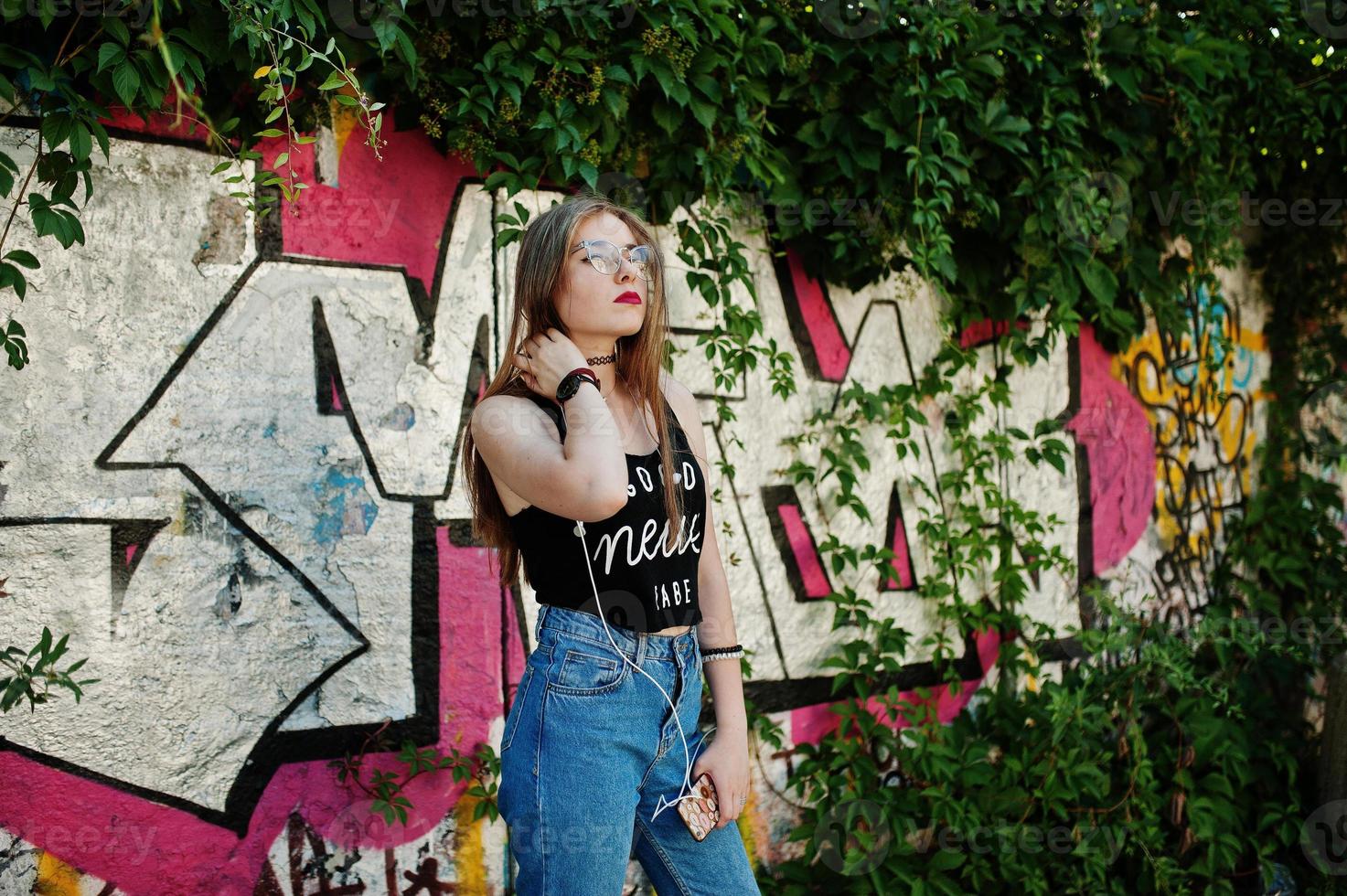 elegante chica hipster casual con gorra, gafas y jeans, escuchando música desde auriculares de teléfono móvil contra una gran pared de graffiti. foto